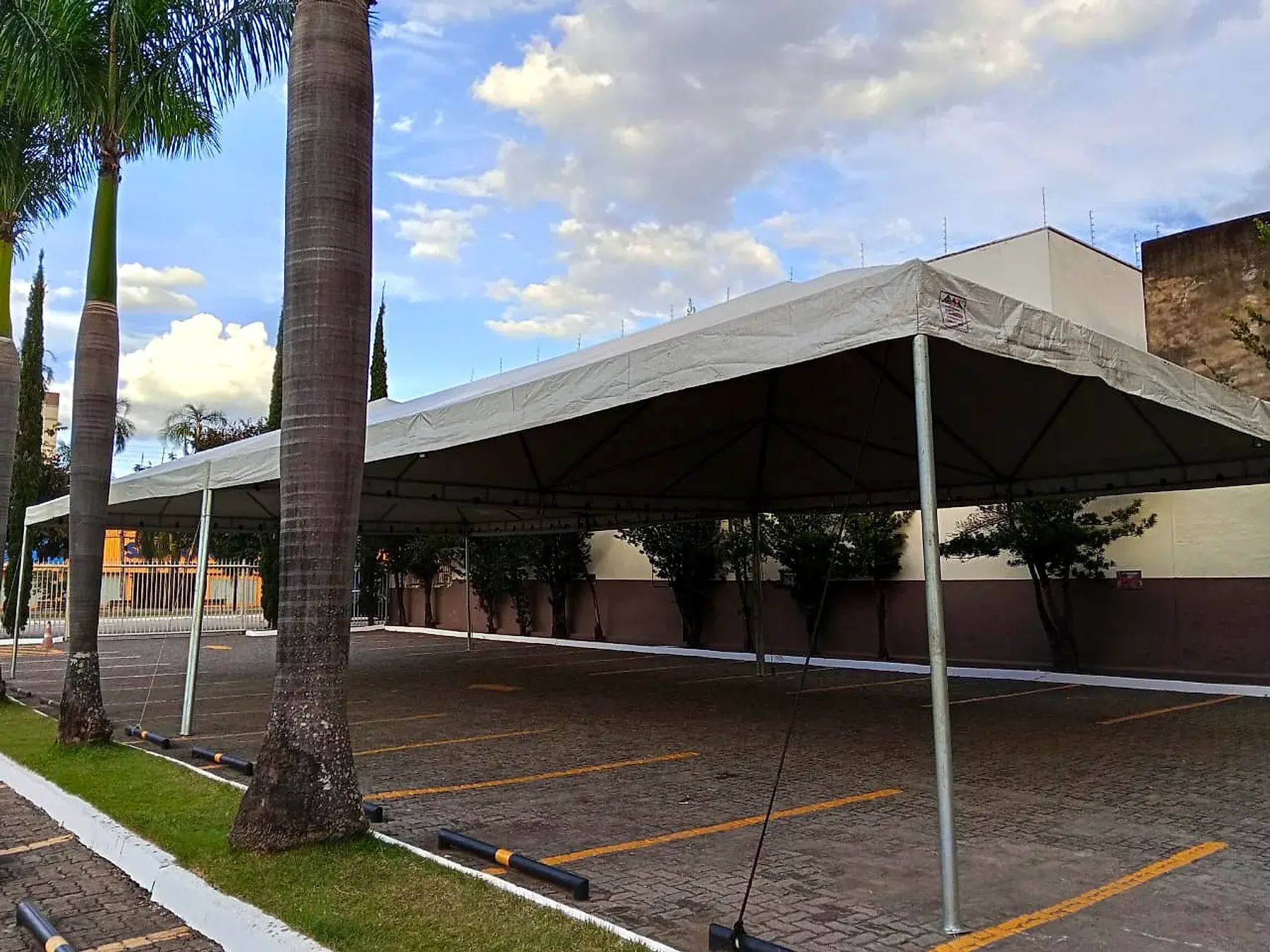 Tenda branca de grande cobertura montada sobre um estacionamento, entre palmeiras, com céu azul ao fundo